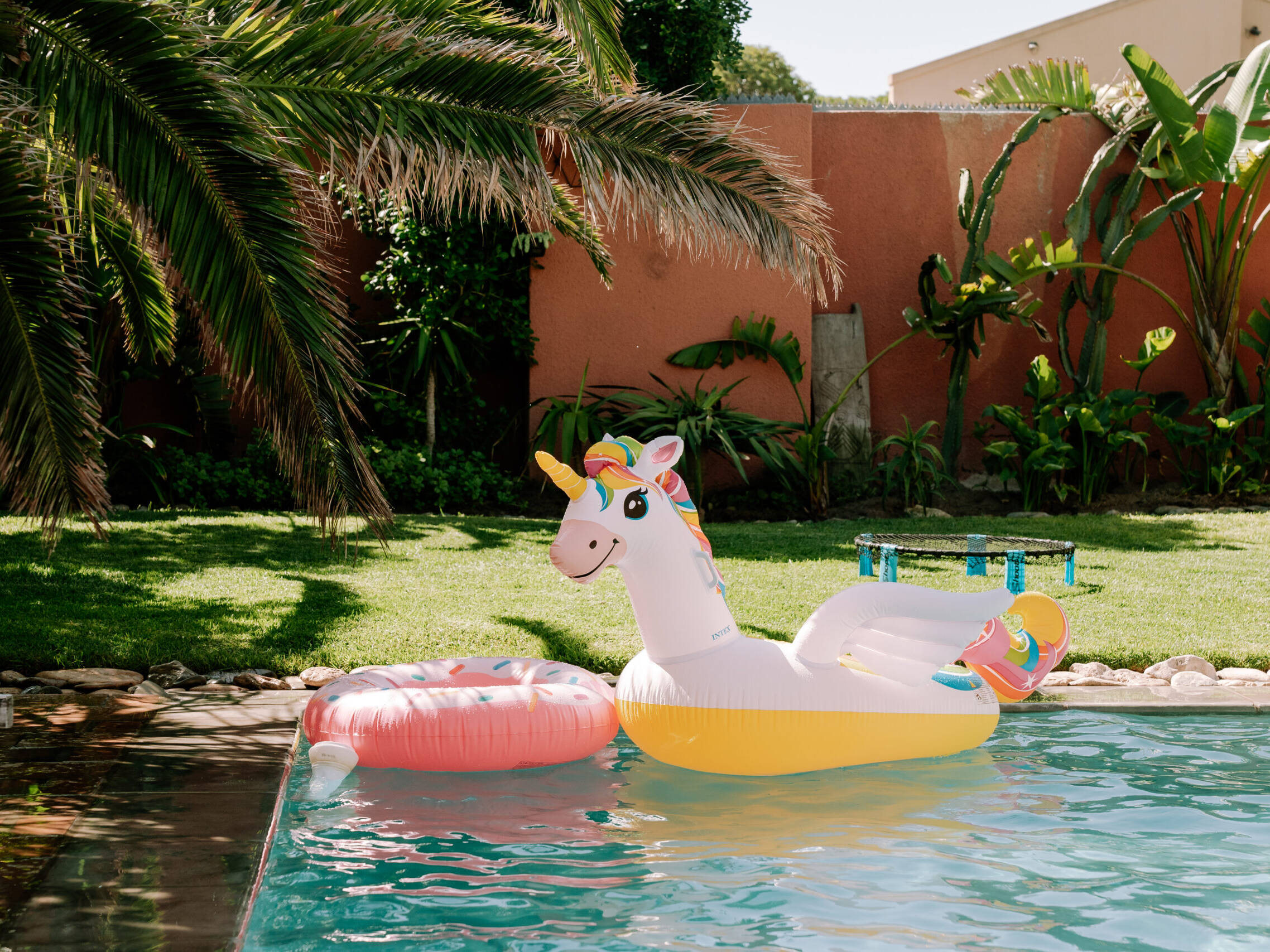 Colorful unicorn and donut inflatable toys floating in the pool at Surfers Lodge, surrounded by lush greenery and palm trees.