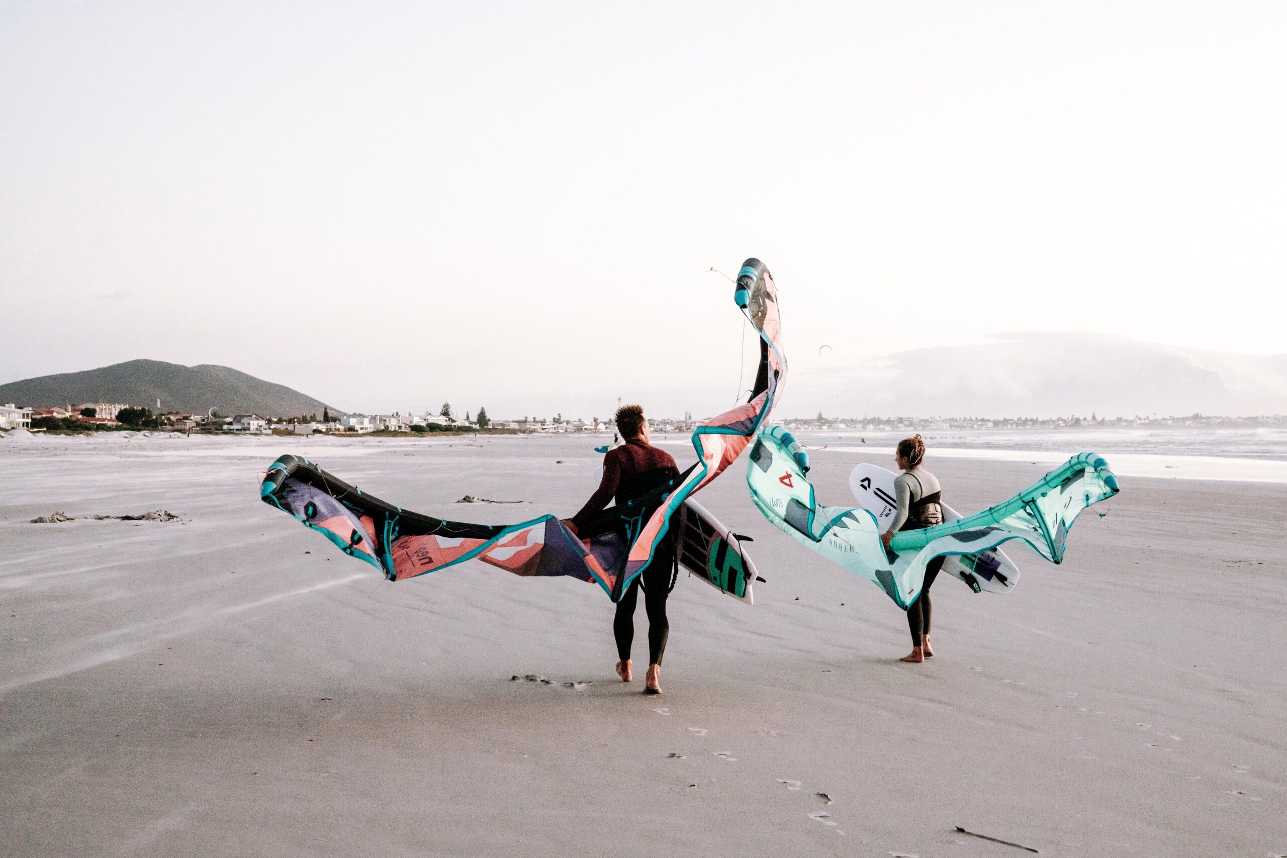 Two young people carrying kitesurfing equipment walking towards the ocean on a beach near Cape Town Surfers Lodge.