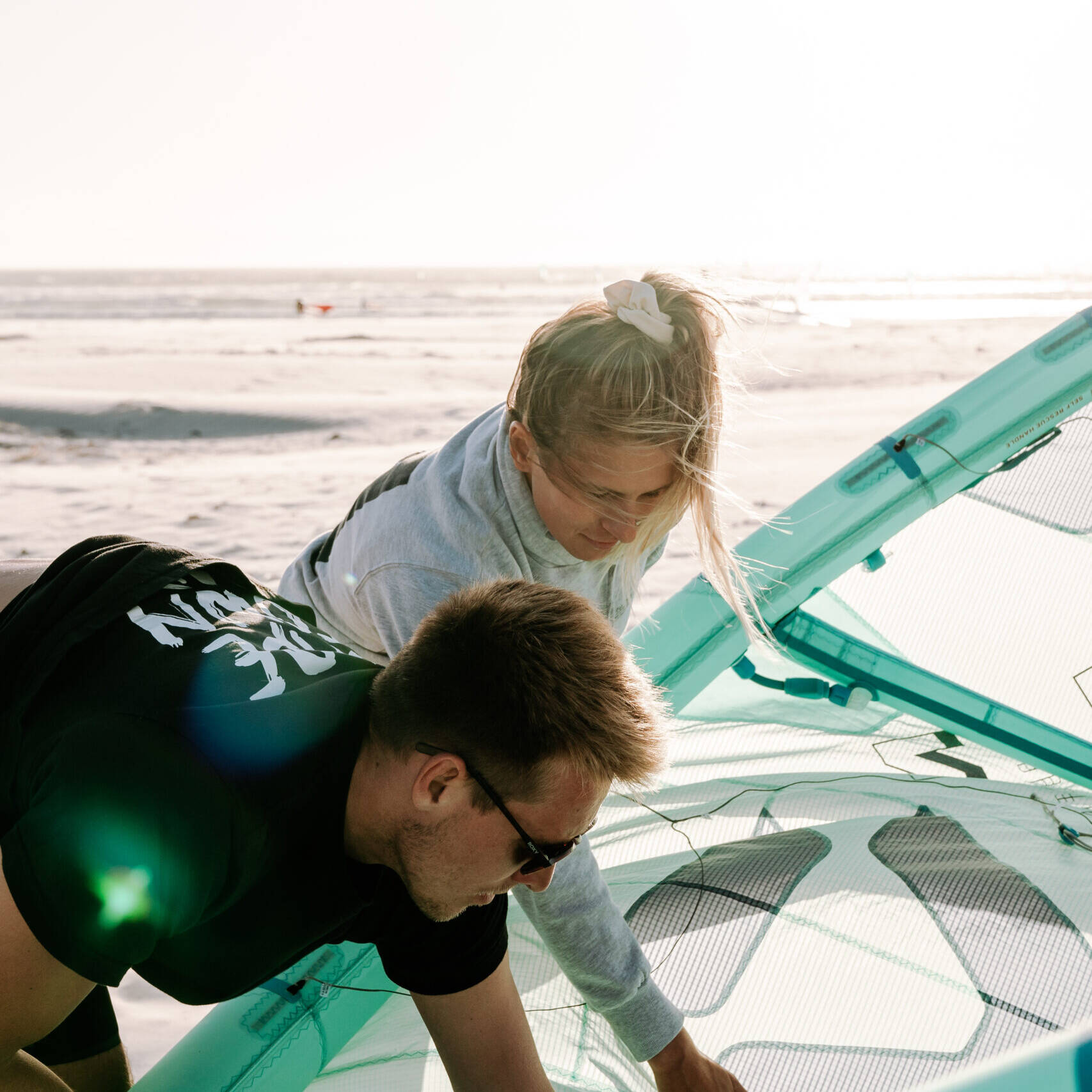 Two people setting up a kitesurfing kite on a beach. A blonde woman in a grey hoodie is leaning forward while a man with sunglasses and a black T-shirt is working on the turquoise kite.