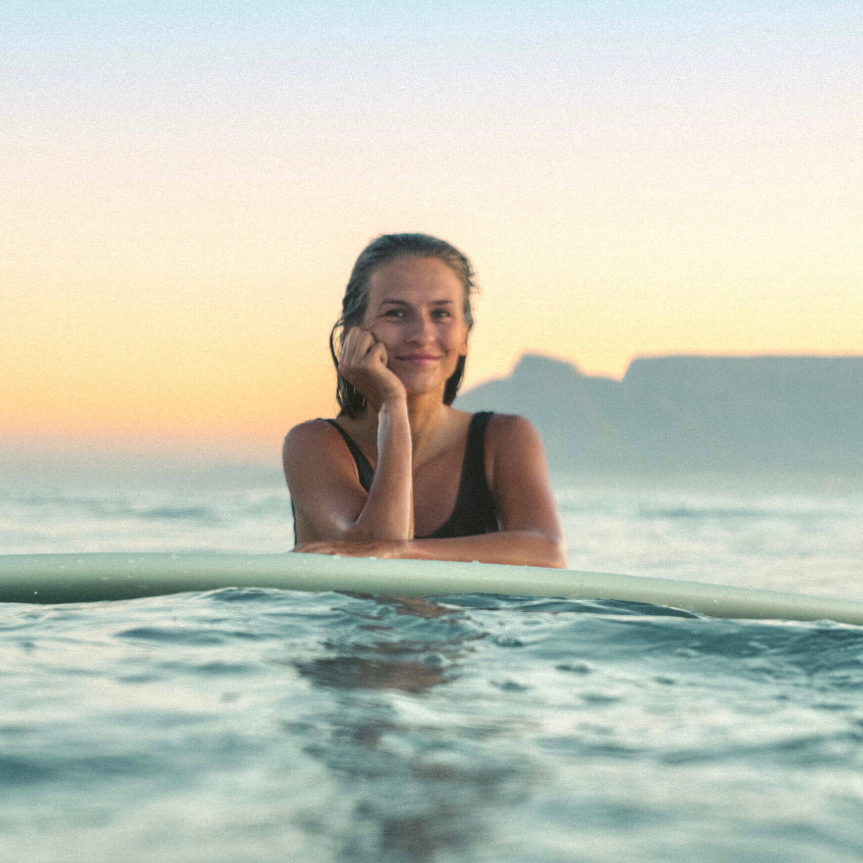 Emely, one of the hosts of Cape Town Lodges, leaning on a surfboard in the ocean during sunset with Table Mountain in the background.