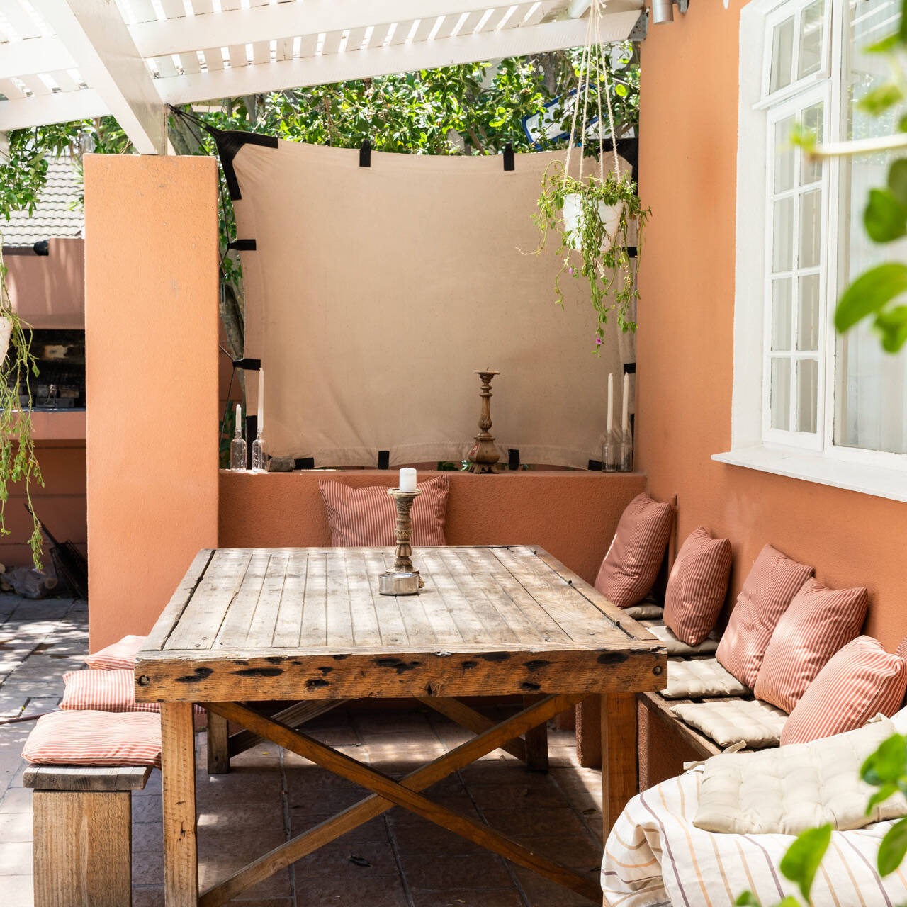 Cozy outdoor dining area with a wooden table, cushioned benches, pink pillows, hanging plant, and built-in grill. The space has a warm terracotta-colored wall and is adorned with candles, creating a cozy and inviting atmosphere.