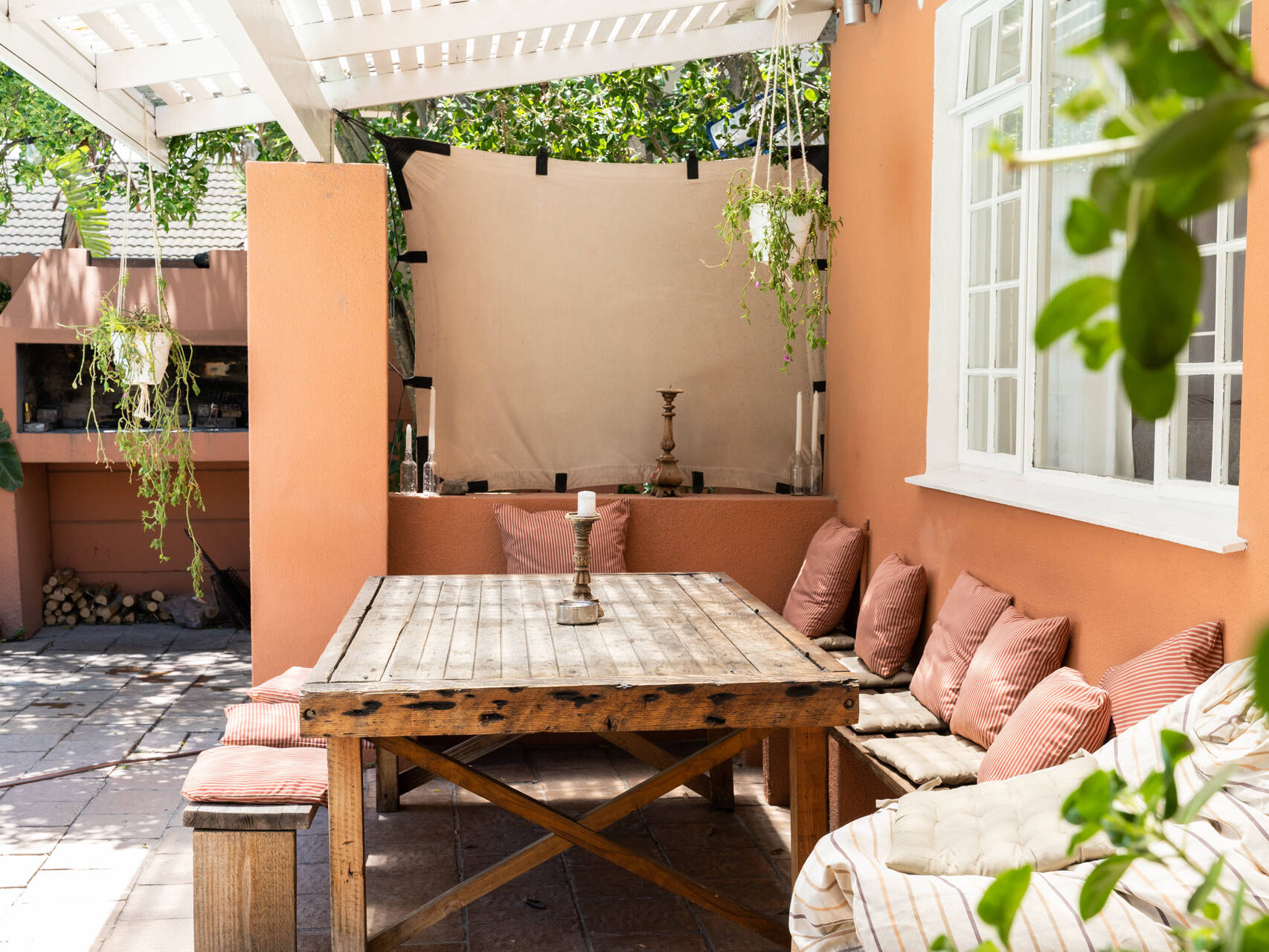 Cozy outdoor dining area with a wooden table, cushioned benches, pink pillows, hanging plant, and built-in grill. The space has a warm terracotta-colored wall and is adorned with candles, creating a cozy and inviting atmosphere.