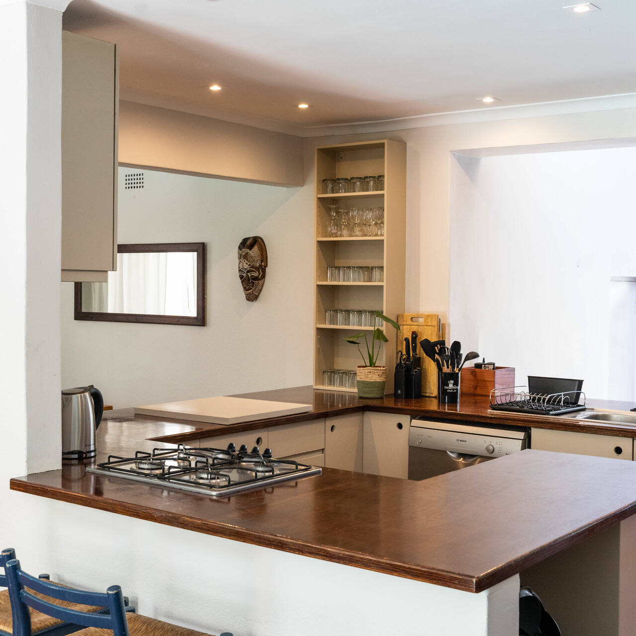 Modern communal kitchen with a wooden countertop, integrated gas stove, dual sink, open shelving with glasses, and a wall-mounted chalkboard schedule. The kitchen includes essential appliances such as a dishwasher and an electric kettle, with decorative touches like a potted plant and a wall mask.