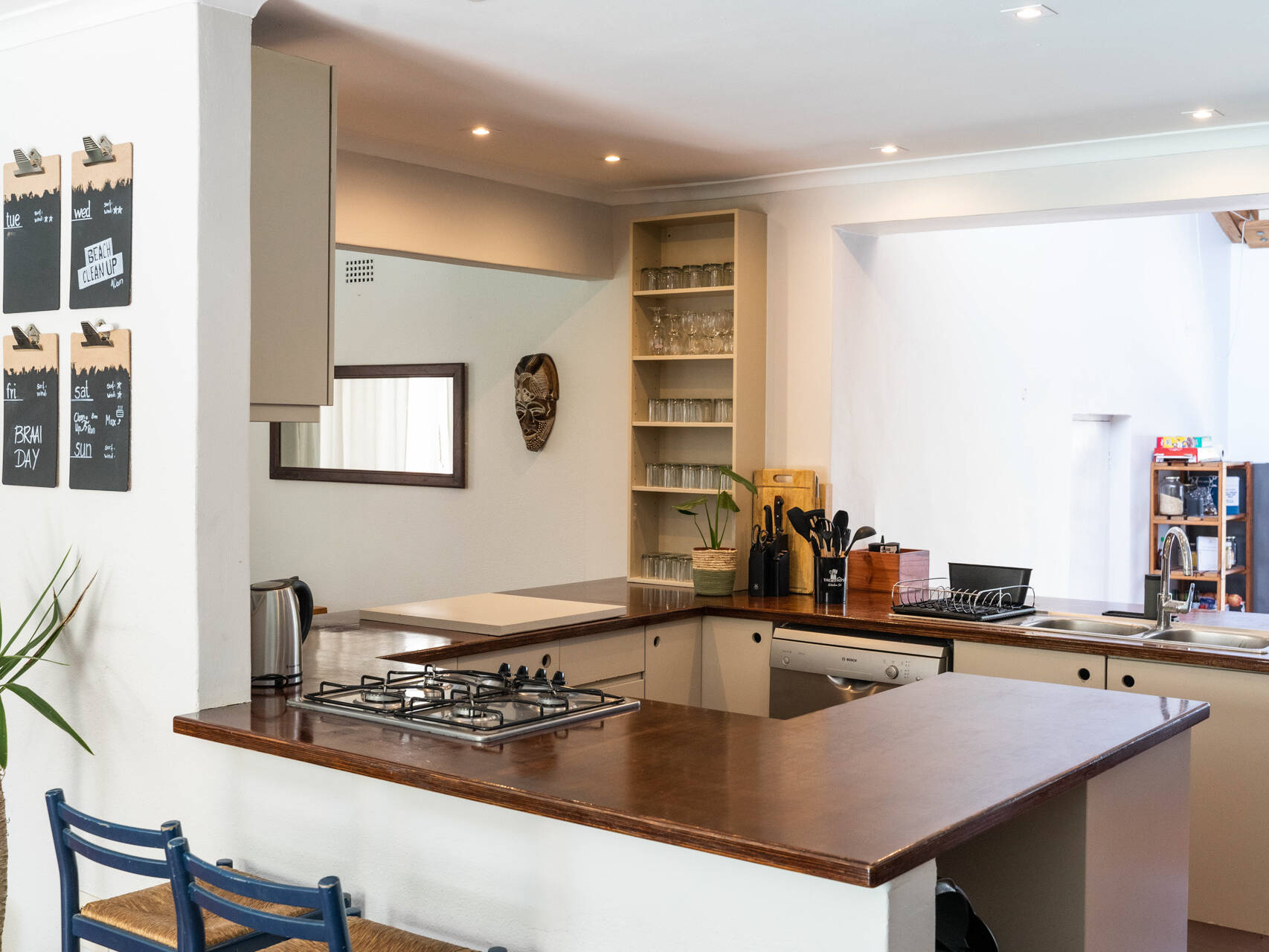 Modern communal kitchen with a wooden countertop, integrated gas stove, dual sink, open shelving with glasses, and a wall-mounted chalkboard schedule. The kitchen includes essential appliances such as a dishwasher and an electric kettle, with decorative touches like a potted plant and a wall mask.