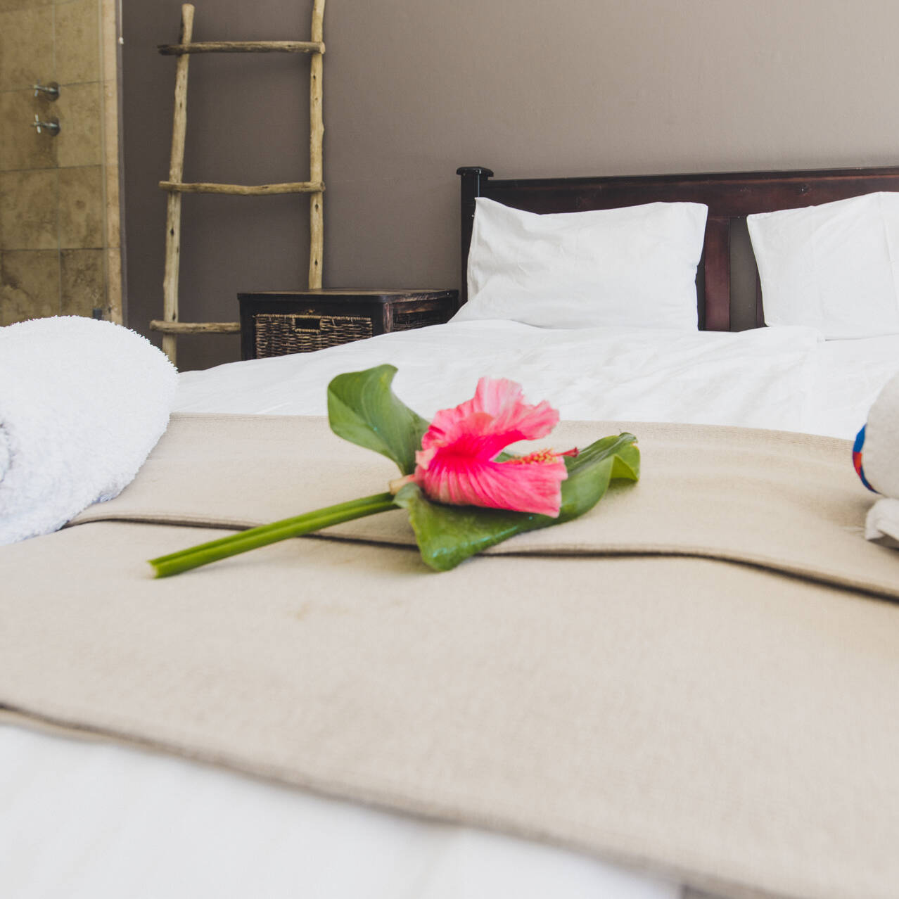 Inviting poolside double room at Cape Town Surfers Lodge with a double bed, white linens, a pink flower on a beige bed runner, rustic wicker bedside tables, a wooden towel ladder, and a tiled bathroom in the background.