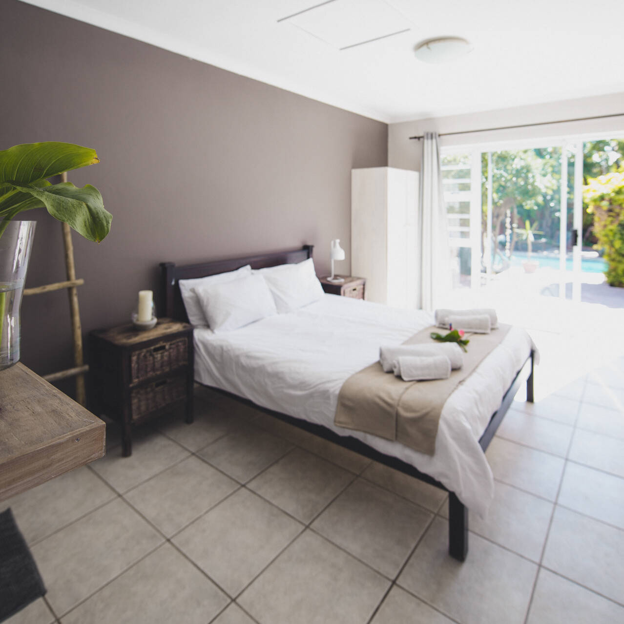 Bright and cozy poolside double room at Cape Town Surfers Lodge with a double bed, rustic wooden furniture, a vase with green leaves, and sliding glass doors opening to a view of the pool.