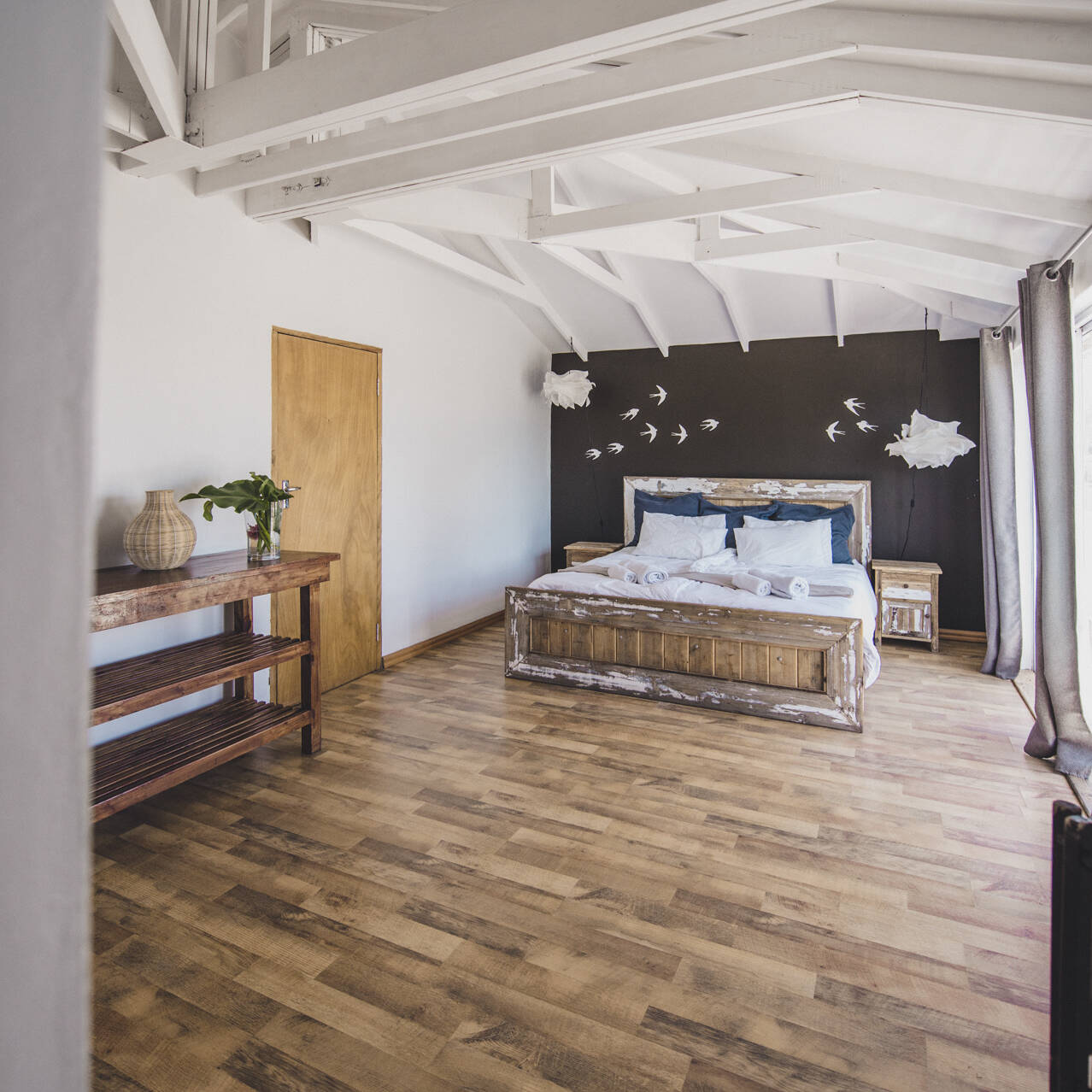 Spacious master bedroom in the poolside apartment at Cape Town Surfers Lodge with a rustic wooden bed, blue accent pillows, bird wall art, and a wooden console table.