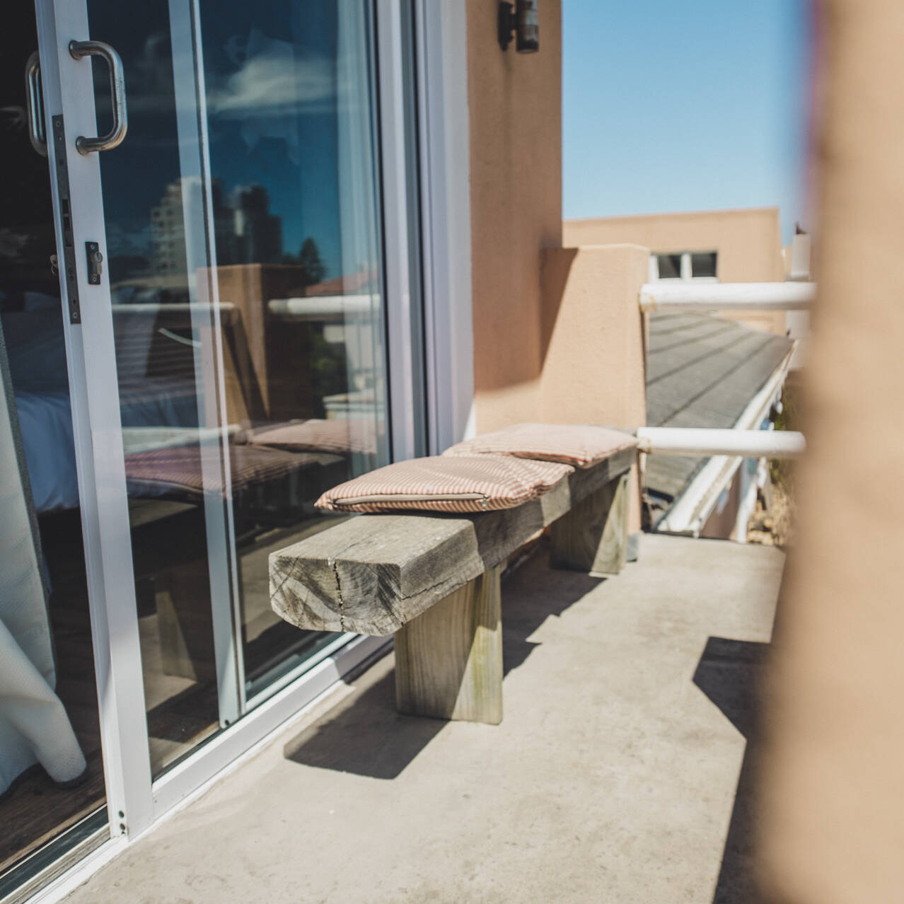 Cozy balcony in the poolside apartment at Cape Town Surfers Lodge with a wooden bench and striped cushions, accessible through sliding glass doors.