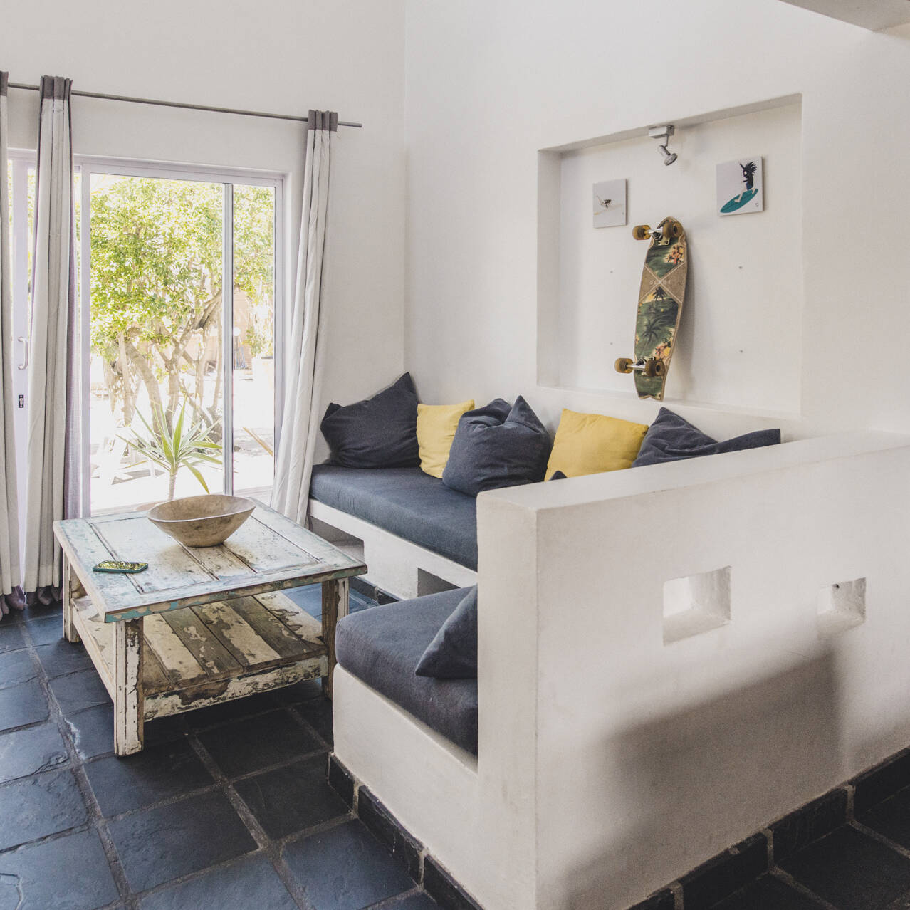 Cozy living area in the poolside apartment at Cape Town Surfers Lodge with built-in seating, a rustic wooden coffee table, skateboard wall decoration, and sliding glass doors opening to a lush patio.