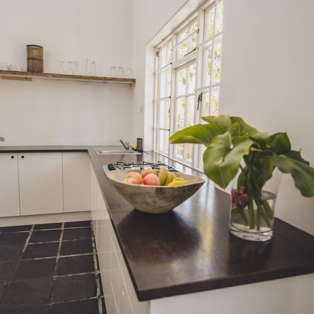 Bright and spacious kitchen in the poolside apartment at Cape Town Surfers Lodge with white cabinetry, a gas stove, a large window, a bowl of fresh fruit, and a vase of green leaves.