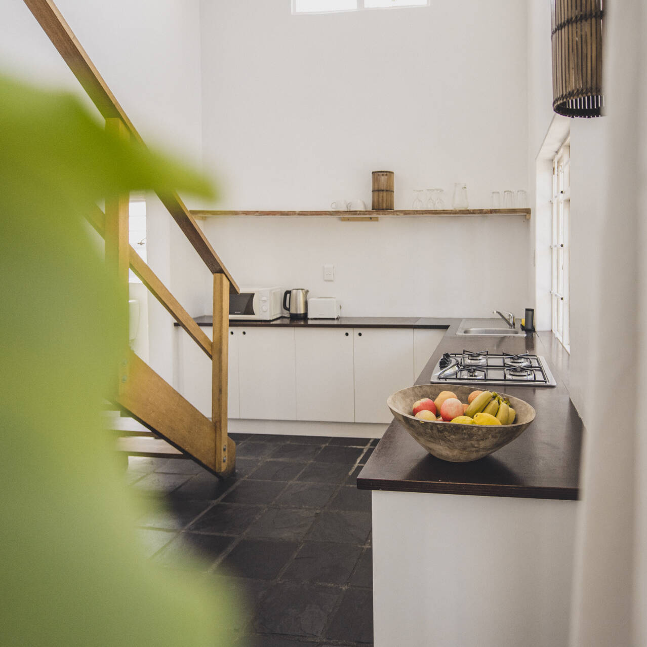 Modern kitchen in the poolside apartment at Cape Town Surfers Lodge with white cabinetry, a gas stove, a wooden staircase, and a bowl of fresh fruit.