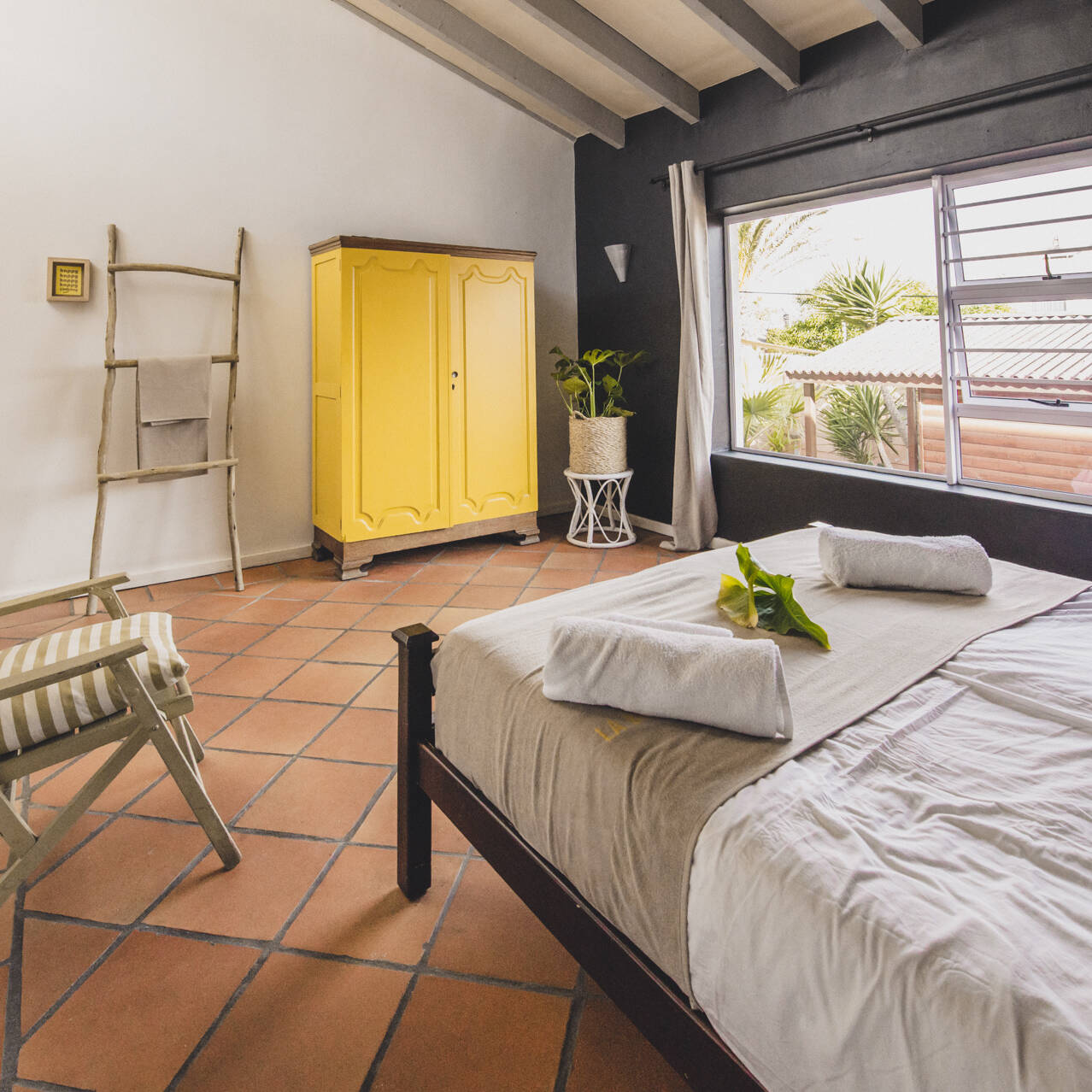 Cozy Premium Double Room at Surfers Lodge with a double bed, yellow wardrobe, rustic ladder-style towel rack, and large window providing natural light.