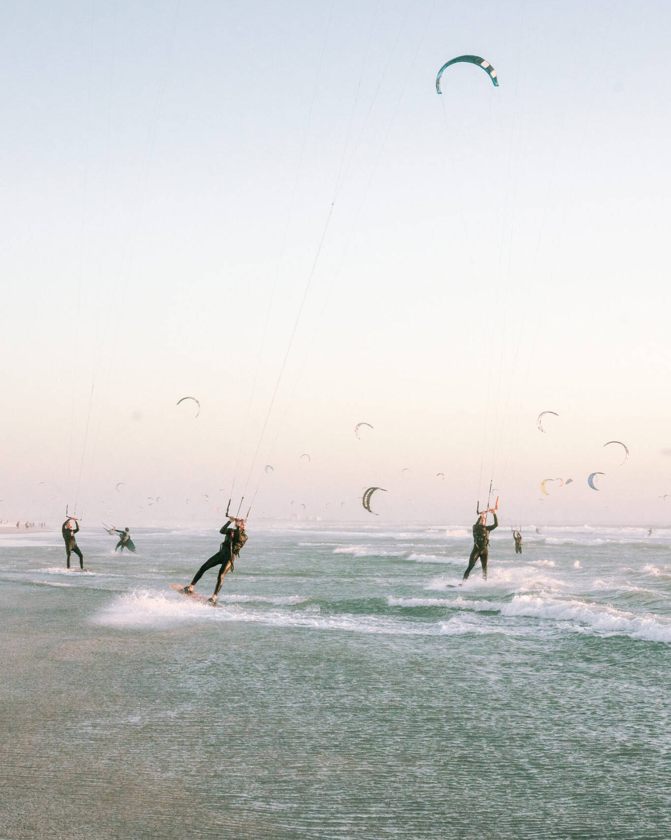 Kitesurfers riding the waves at Kite Beach in Cape Town, with numerous colorful kites in the sky and people walking along the shore.