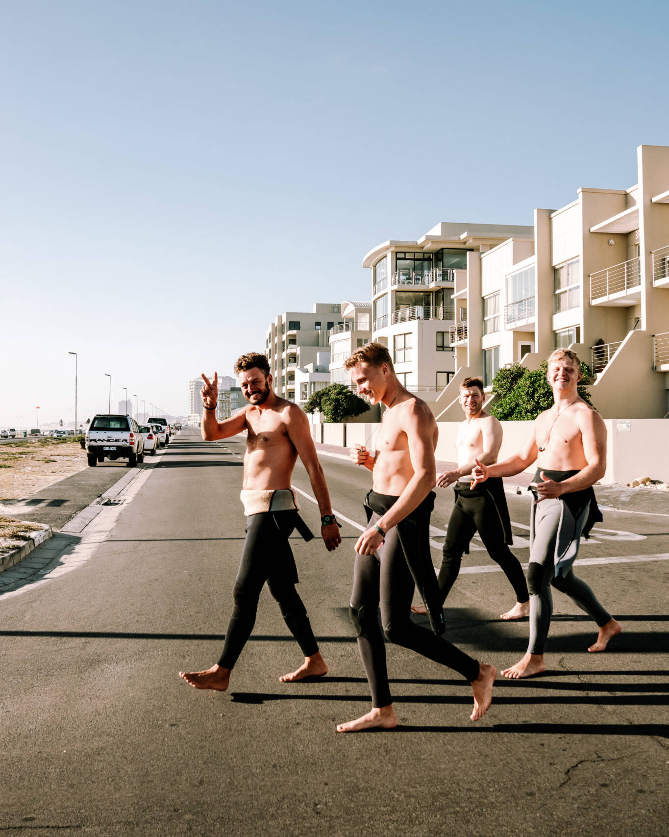 Kitesurfers walking from the Surfer's Lodge and Surf Hostel to Kite Beach, wearing wetsuits and carrying their gear, with beachfront apartments and the ocean in the background.