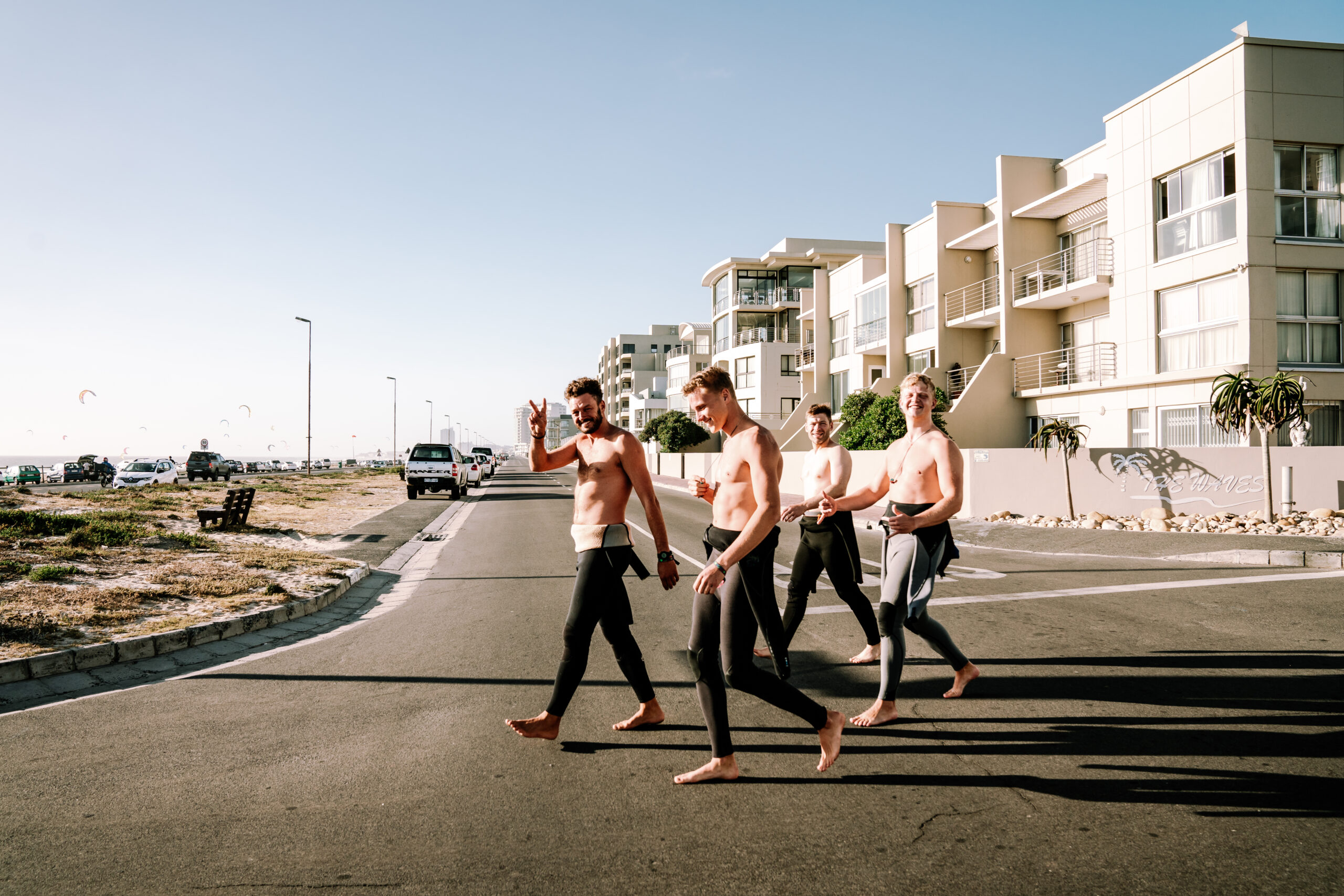 Kitesurfers walking from the Surfer's Lodge and Surf Hostel to Kite Beach, wearing wetsuits and carrying their gear, with beachfront apartments and the ocean in the background.