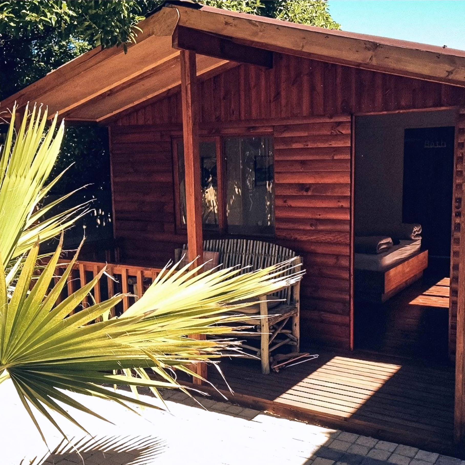 Charming Surf Shack at Cape Town Surfers Lodge featuring a rustic wooden exterior, a cozy porch with a wicker chair, and lush greenery including a large palm frond. The interior shows a neatly made bed with plush towels, offering a comfortable and inviting retreat for surfers and nature enthusiasts.