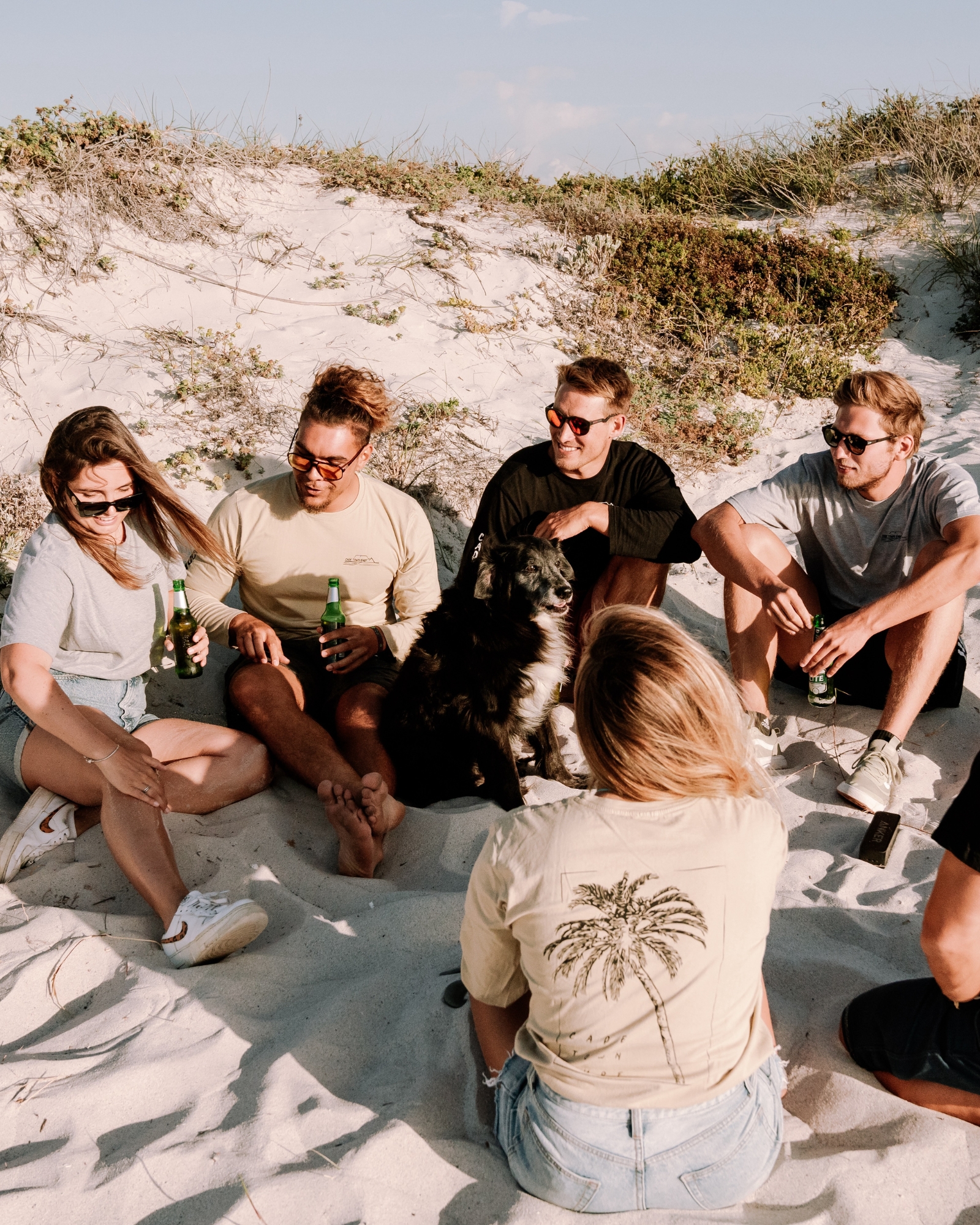 A group of friends sitting on the sand at Blouberg beach, enjoying drinks and the company of a friendly dog, with dunes and greenery in the background.