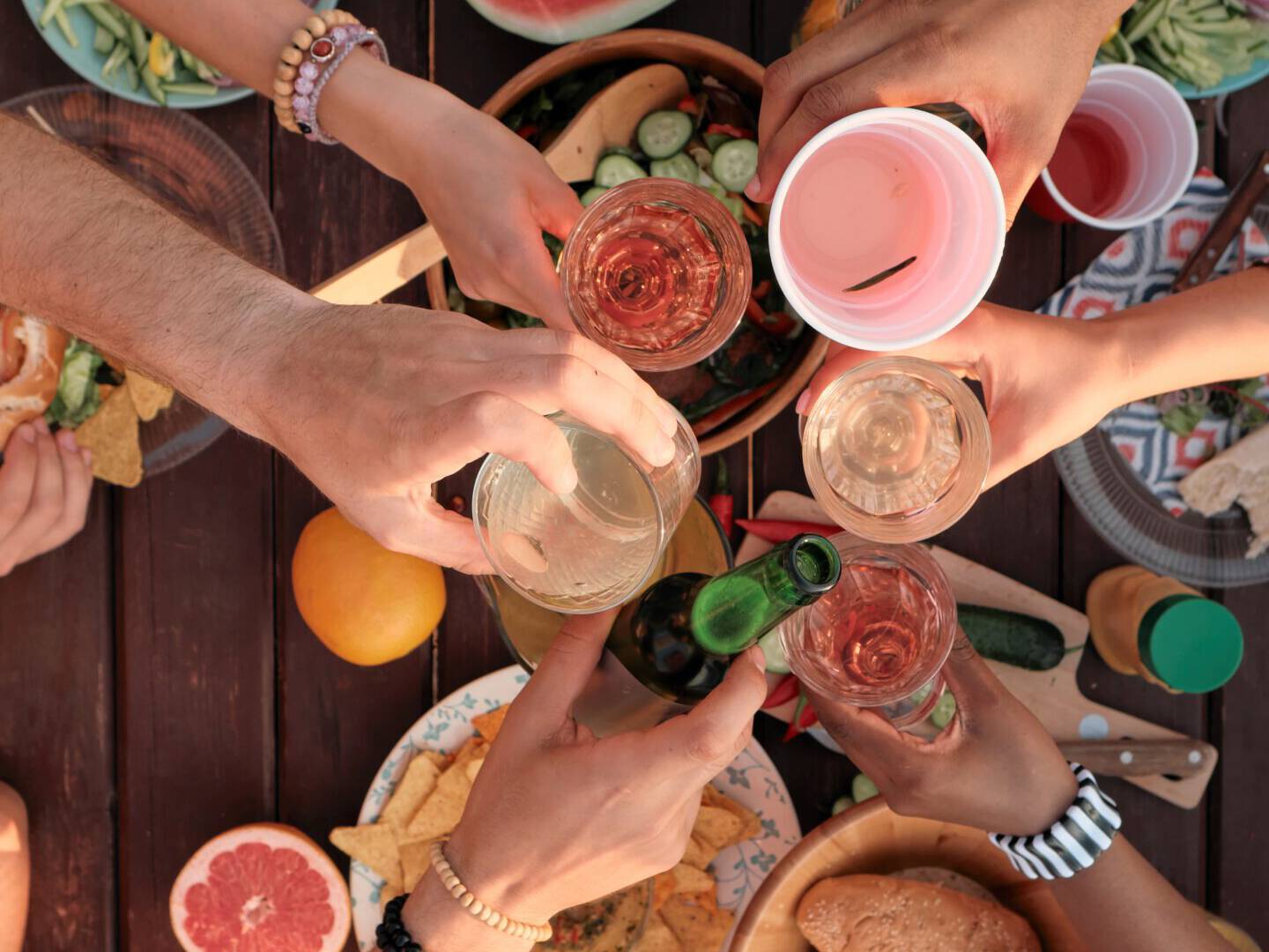 A group of friends toasting over a table filled with a variety of fresh and colorful foods, including watermelon slices, skewers, assorted breads, and fruits at Cape Town Surfers Lodge. The outdoor feast scene is vibrant and festive, showcasing the joy of sharing a meal in good company.