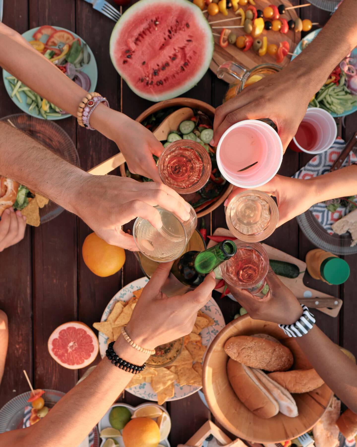 A group of friends toasting over a table filled with a variety of fresh and colorful foods, including watermelon slices, skewers, assorted breads, and fruits at Cape Town Surfers Lodge. The outdoor feast scene is vibrant and festive, showcasing the joy of sharing a meal in good company.