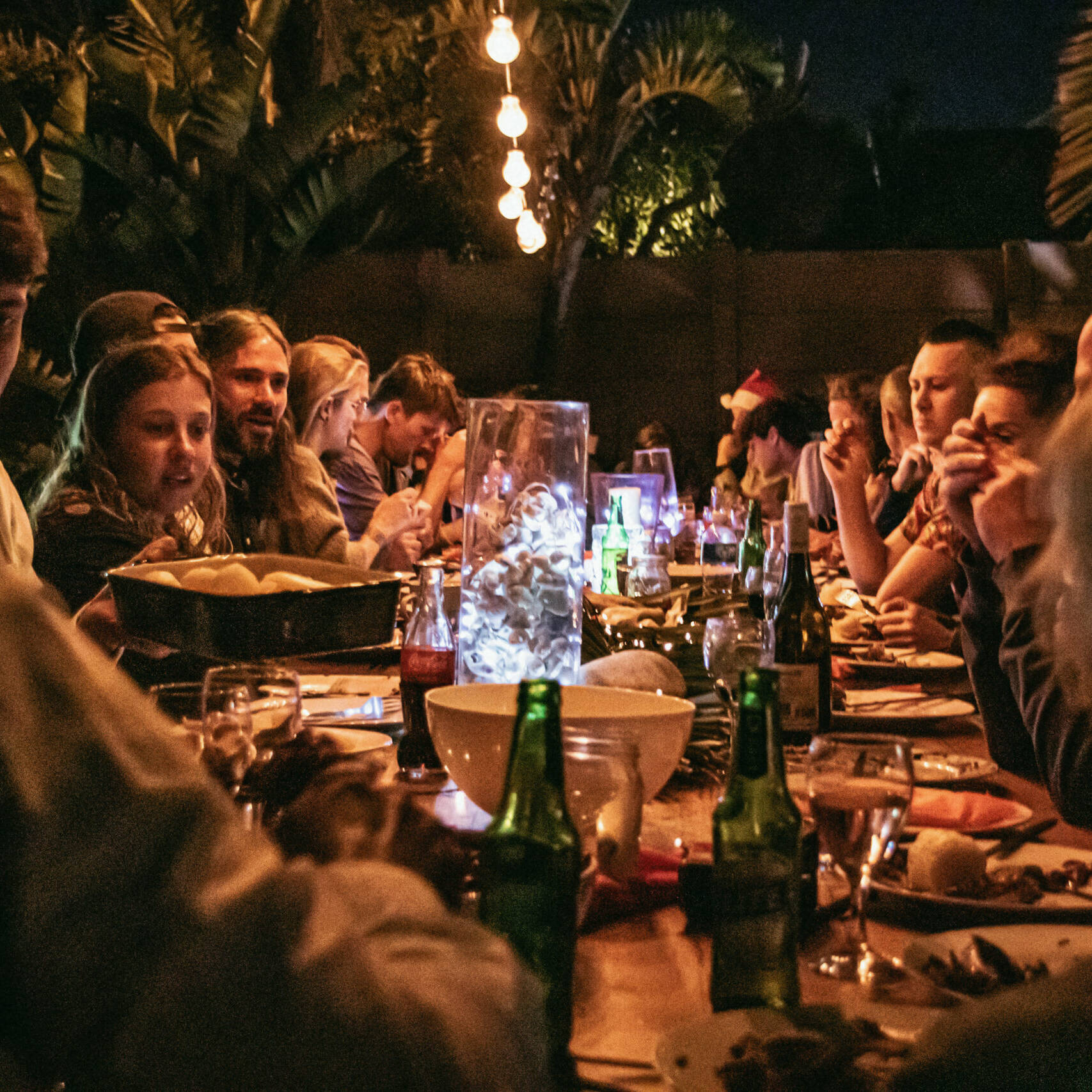 Outdoor communal dinner at night with a long table surrounded by guests enjoying food and drinks under warm hanging lights, creating a lively and festive atmosphere.