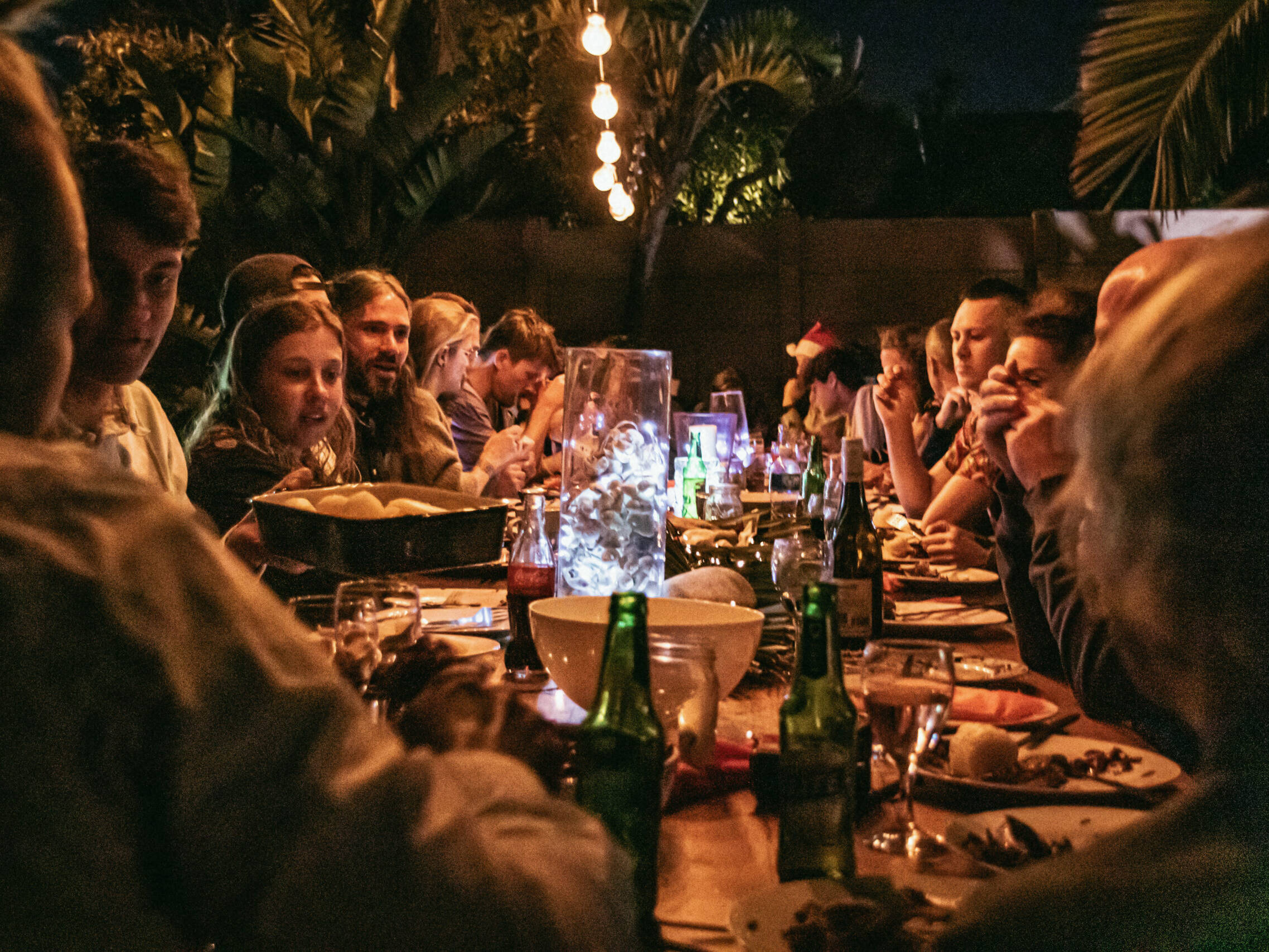 Outdoor communal dinner at night with a long table surrounded by guests enjoying food and drinks under warm hanging lights, creating a lively and festive atmosphere.