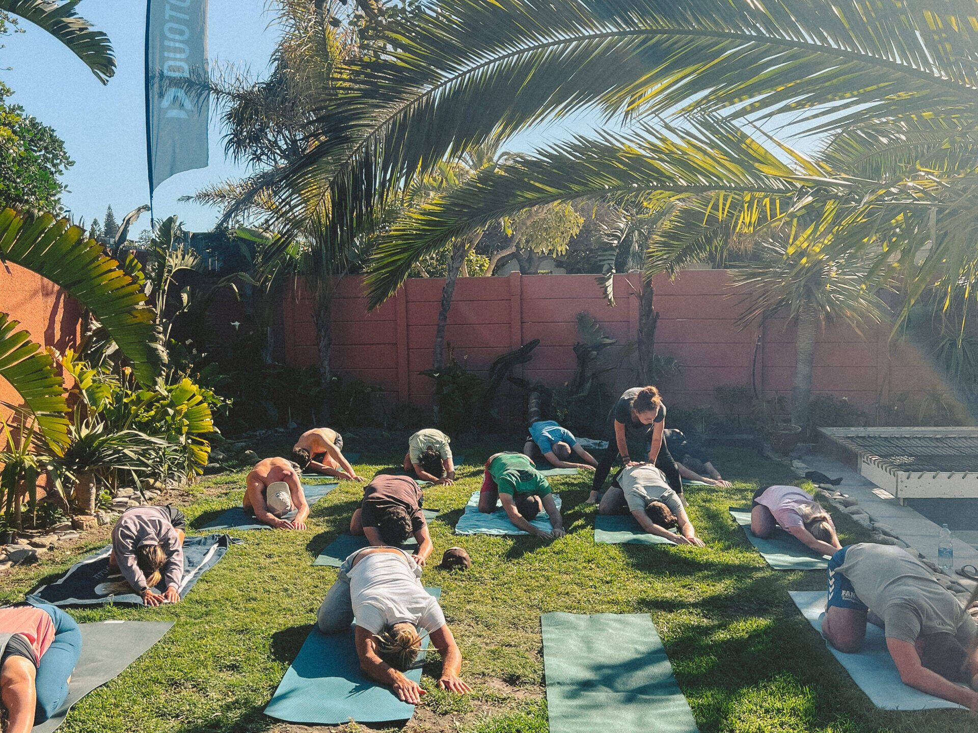 A group of participants engaging in an outdoor yoga session at Cape Town Surfers Lodge, positioned on yoga mats spread across a lush green lawn. Palm trees and a clear blue sky provide a tranquil backdrop, creating an ideal environment for relaxation and rejuvenation.