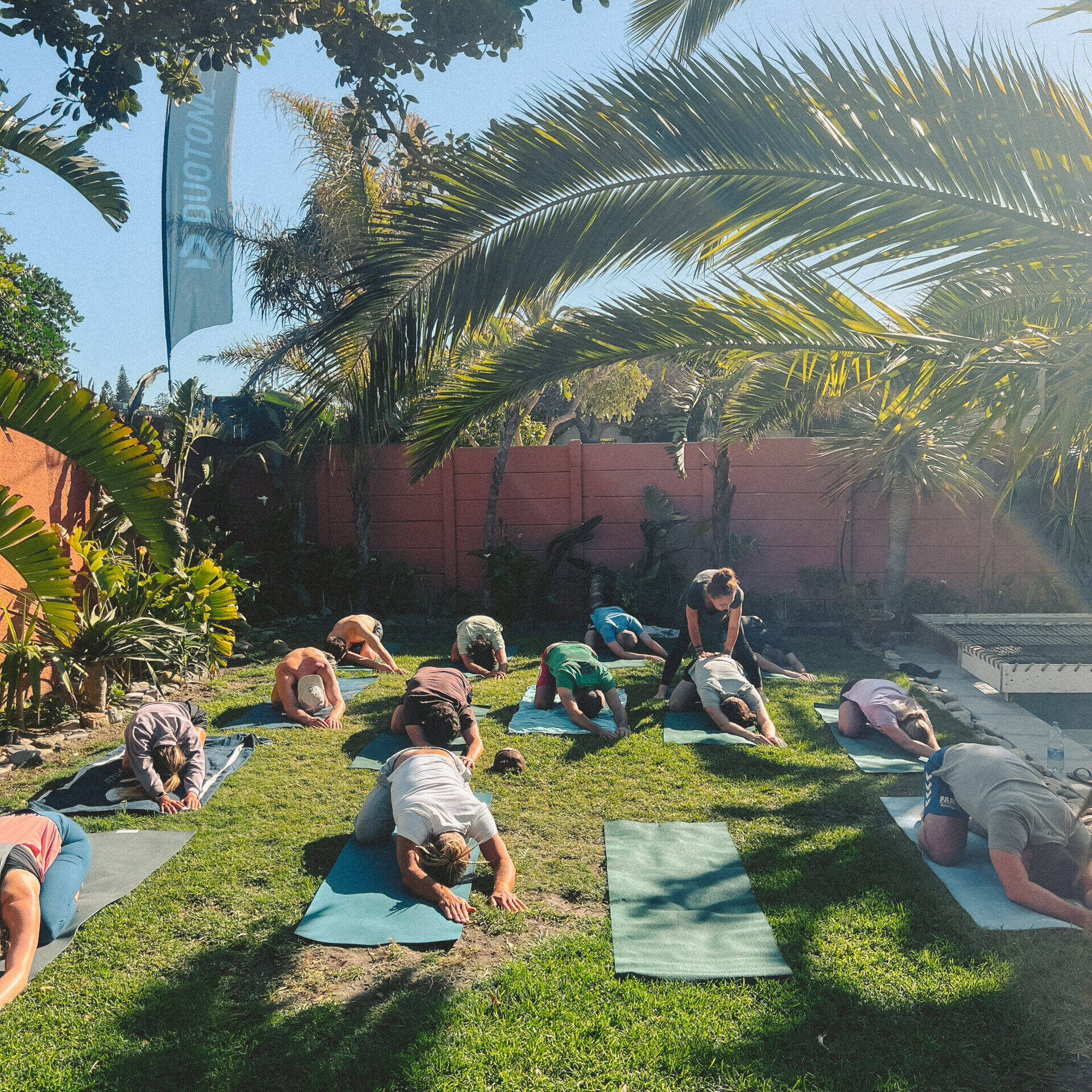 A group of participants engaging in an outdoor yoga session at Cape Town Surfers Lodge, positioned on yoga mats spread across a lush green lawn. Palm trees and a clear blue sky provide a tranquil backdrop, creating an ideal environment for relaxation and rejuvenation.