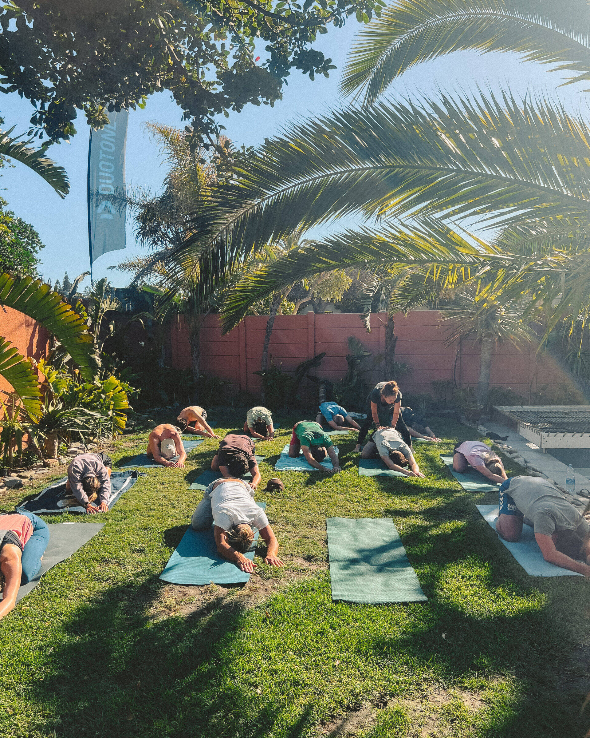 A group of participants engaging in an outdoor yoga session at Cape Town Surfers Lodge, positioned on yoga mats spread across a lush green lawn. Palm trees and a clear blue sky provide a tranquil backdrop, creating an ideal environment for relaxation and rejuvenation.