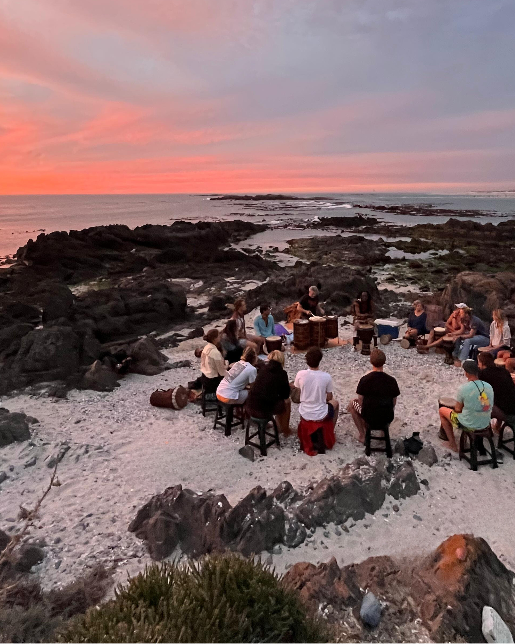 Guests playing djembe drums together at sunset on a rocky Blouberg beach